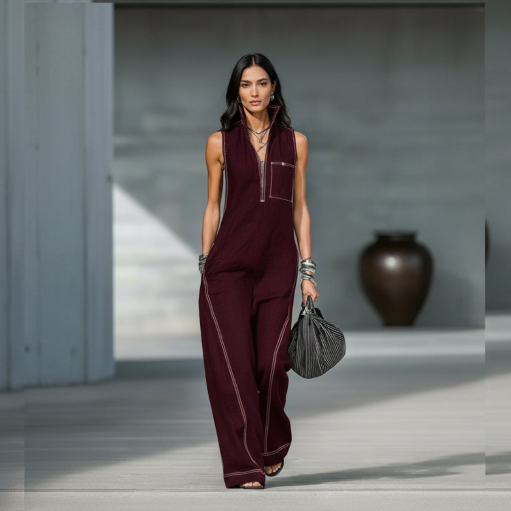 Person wearing a burgundy jumpsuit with white piping, holding a black handbag indoors.