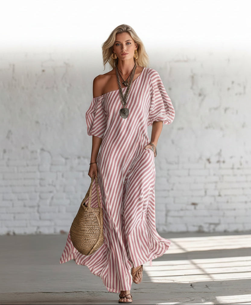 Woman wearing a pink and white striped dress holding a woven bag against a white brick wall.