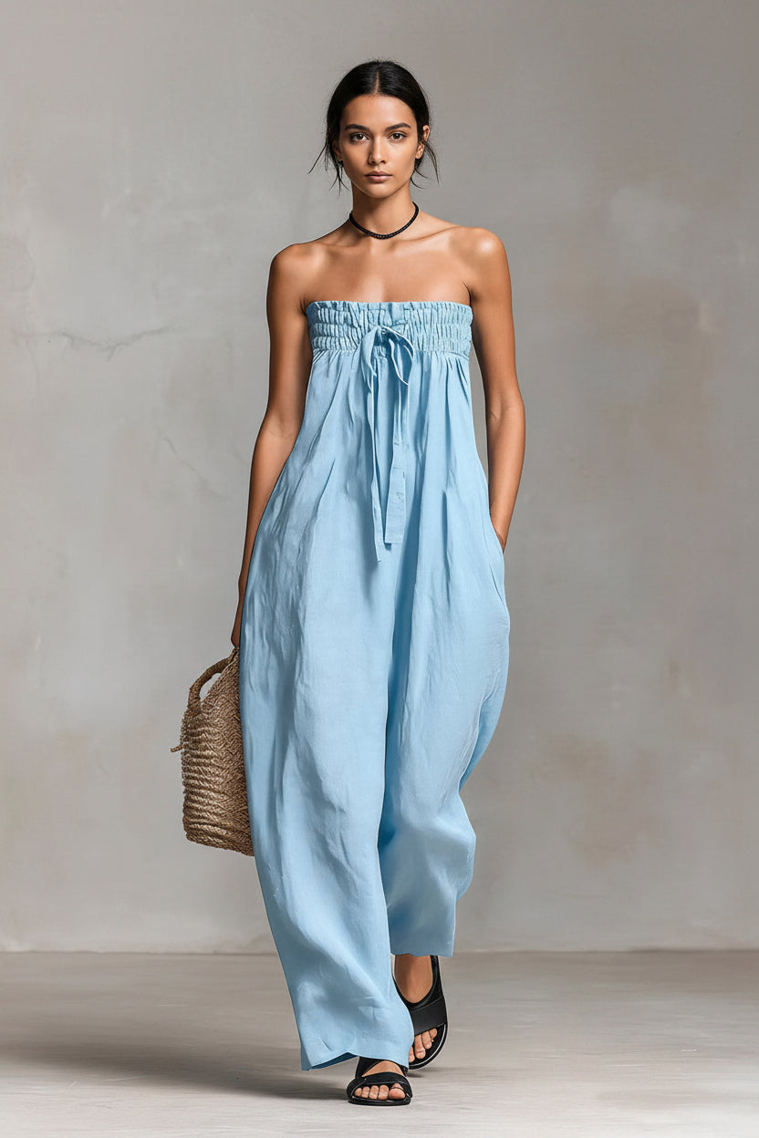 Woman wearing a light blue strapless jumpsuit holding a woven bag against a neutral background