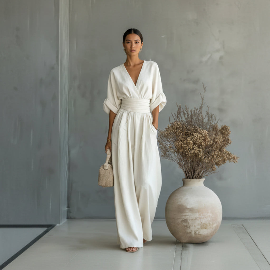Woman in a white dress standing next to a vase with dried flowers against a gray wall.