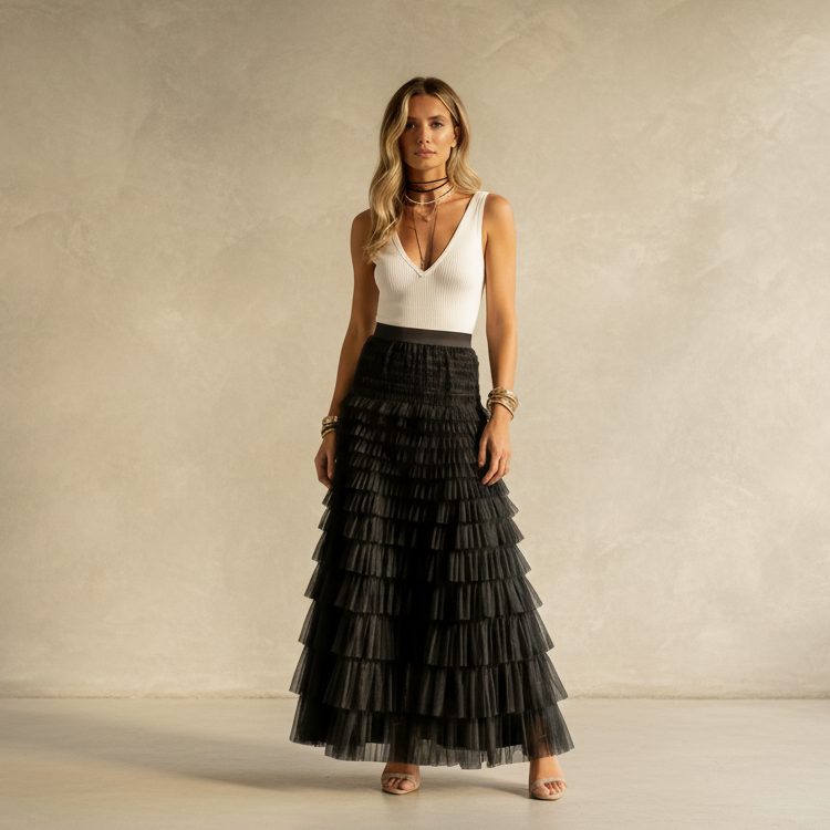 A woman stands in a studio wearing a sleeveless white V-neck top and a long black tiered tulle skirt with a wide black waistband. She is posed against a textured beige wall with a light floor.