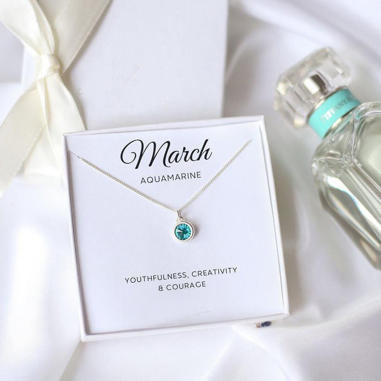 Aquamarine necklace on a card with 'March' and 'Aquamarine' text, next to a bottle of perfume on a white satin background.