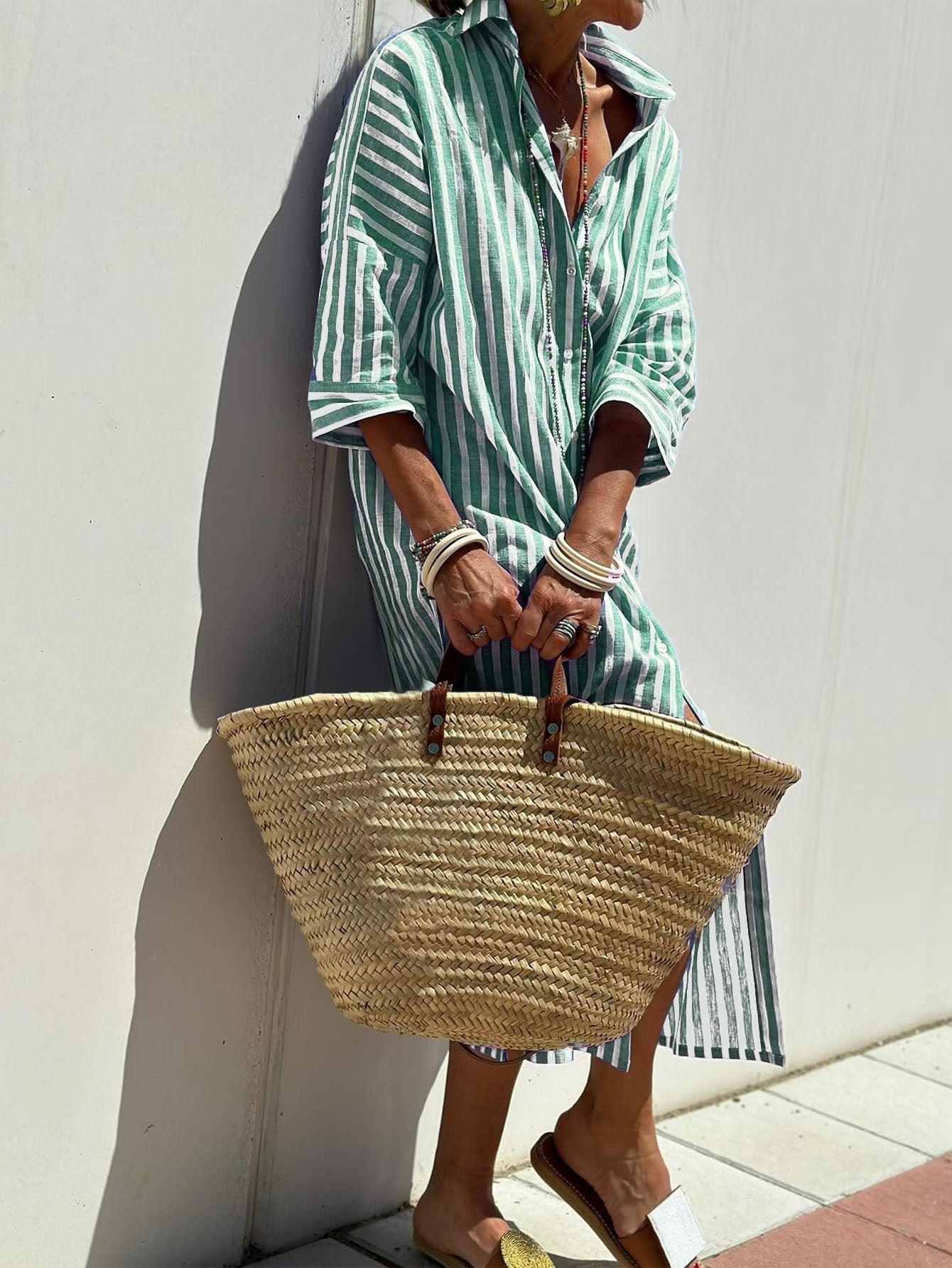 Person wearing a green and white striped dress holding a straw bag against a white wall.