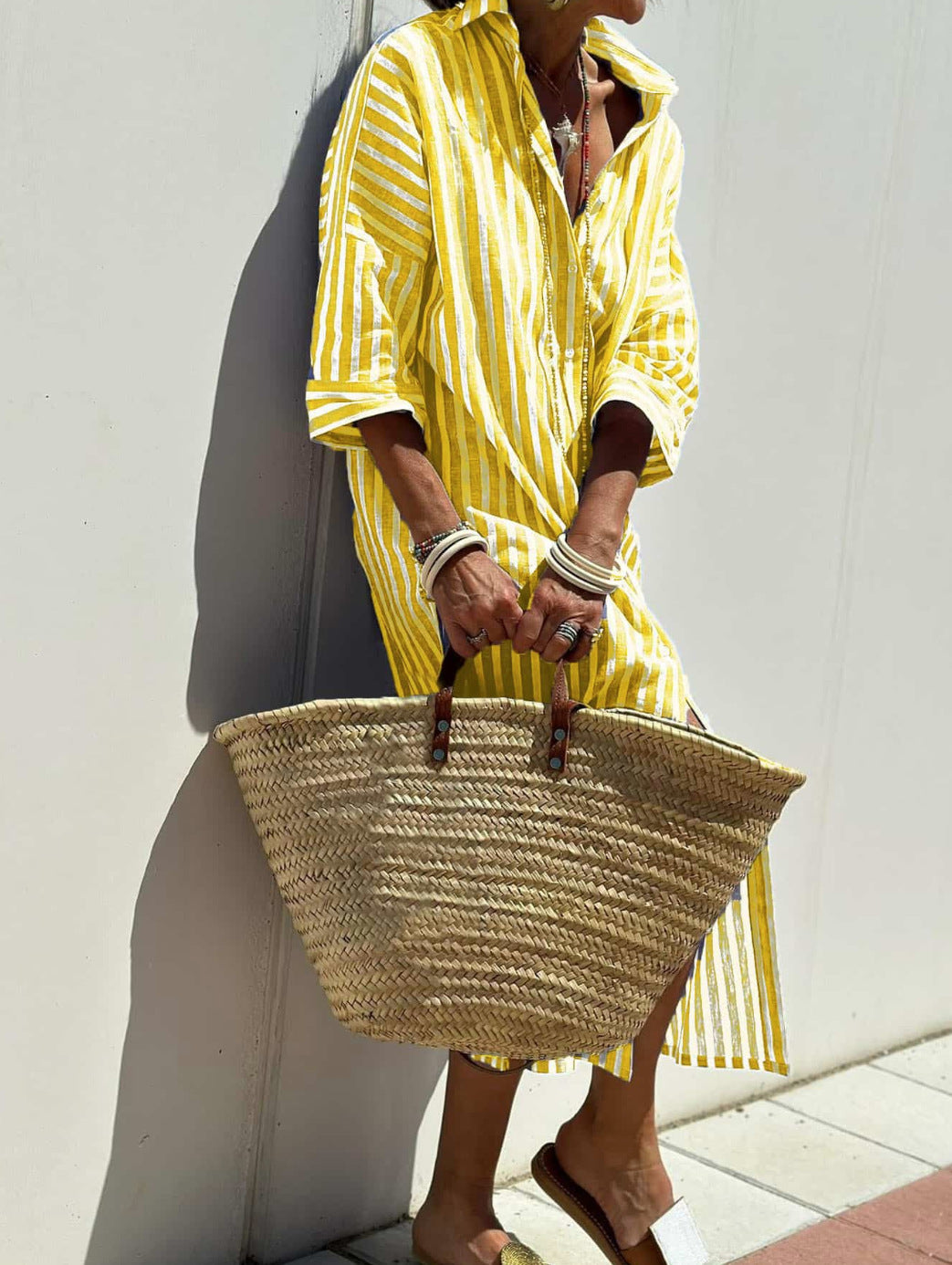 Person wearing a yellow striped dress holding a straw bag against a white wall.