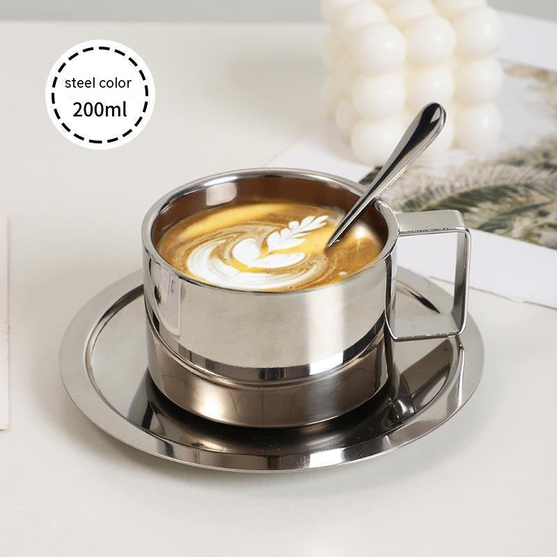 Stainless steel cup with latte art and spoon on a matching saucer, on a light background.