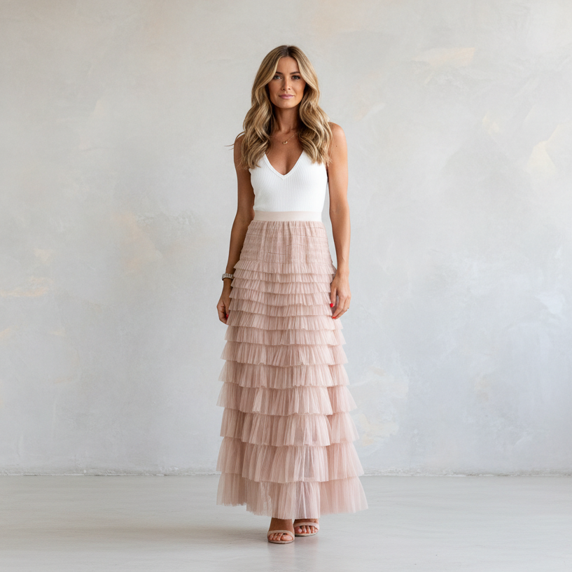 A woman stands in a studio wearing a sleeveless white V-neck top and a long, tiered blush pink tulle maxi skirt, paired with brown heeled sandals against a light textured wall.