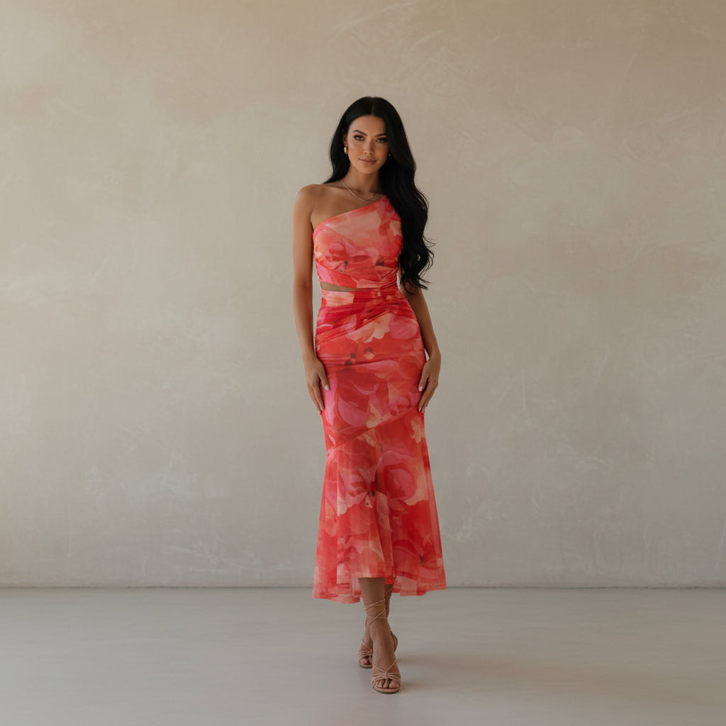 Woman wearing a pink and red one-shoulder dress against a concrete wall with a vase of dried plants.